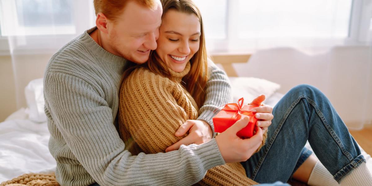 Happy couple sitting on a bed at home, exchanging a small red gift and smiling